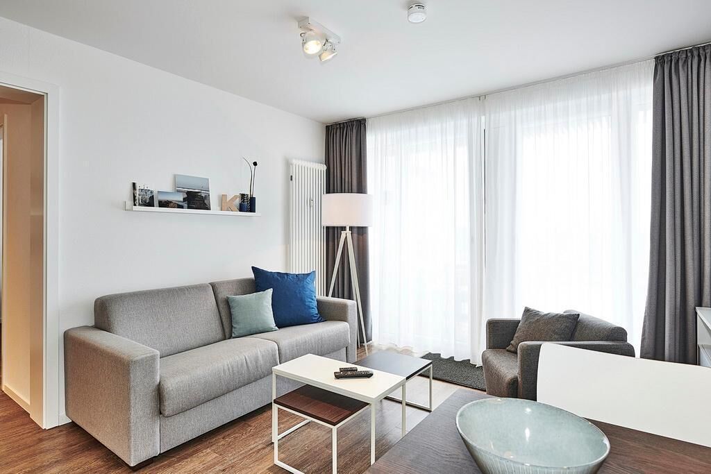 A modern living room with a grey sofa, a coffee table, and a floor lamp. There is a window with curtains and a radiator on the wall.