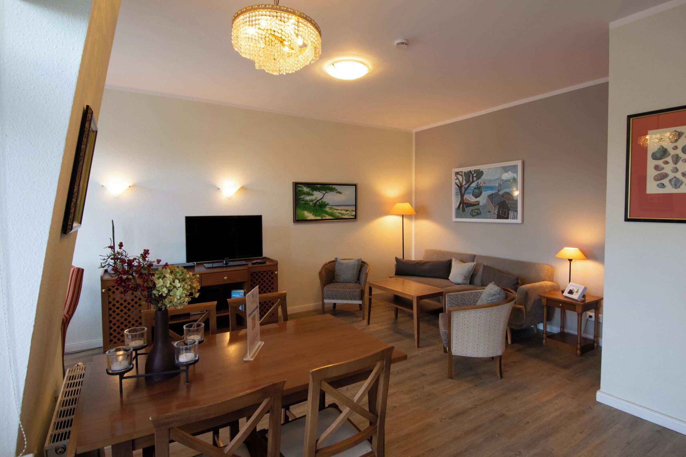A living room with a wooden dining table, chairs, a TV, and a sofa. There are wall-mounted lamps and framed artwork.