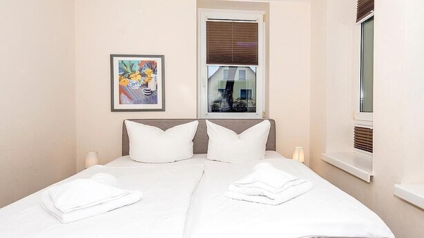 Un lit double avec du linge de lit blanc et des oreillers, une table de chevet avec une lampe, une fenêtre avec des stores et une œuvre d’art encadrée au mur.