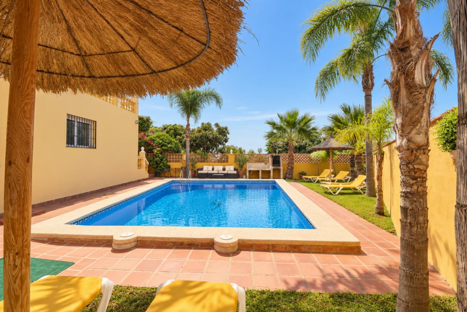Une piscine dotée d’une terrasse carrelée, entourée de palmiers et agrémentée d’un parasol en chaume.