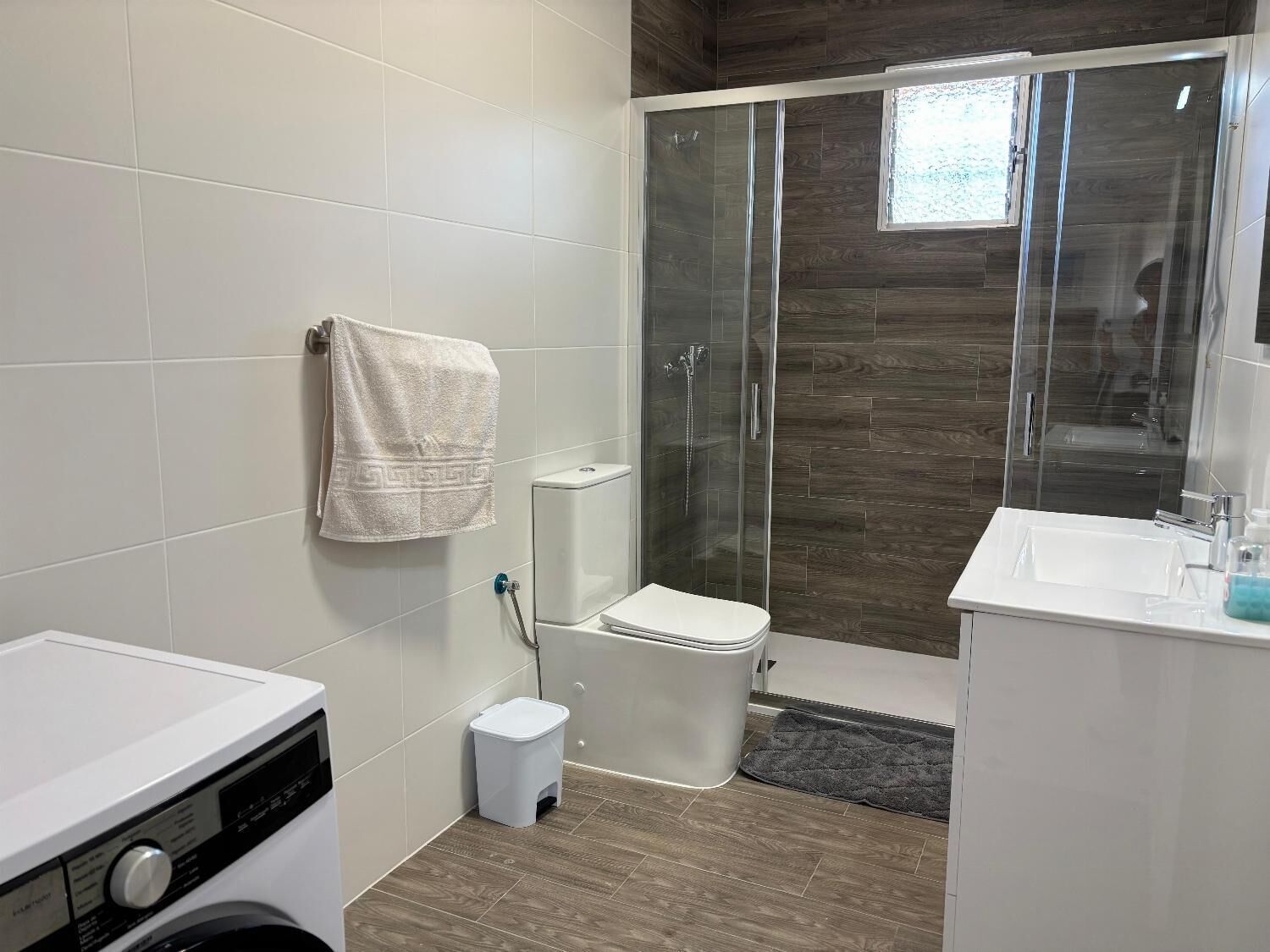 Et moderne badeværelse med en glasafskærmet brusekabine, toilet, vaskemaskine og et håndvaskebord med toiletartikler.