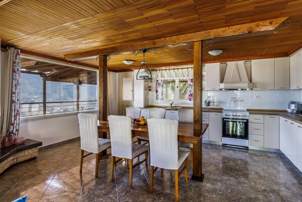 A dining area with a wooden table and chairs, a kitchen with white cabinets, and a view through a window. - Villa Aloupi - Three Bedroom Villa, Sleeps 6 (Sporades)