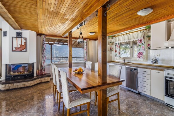 Amplia sala de estar con una mesa de comedor de madera, sillas, chimenea y cocina con armarios blancos y ventana con vista a un paisaje montañoso.