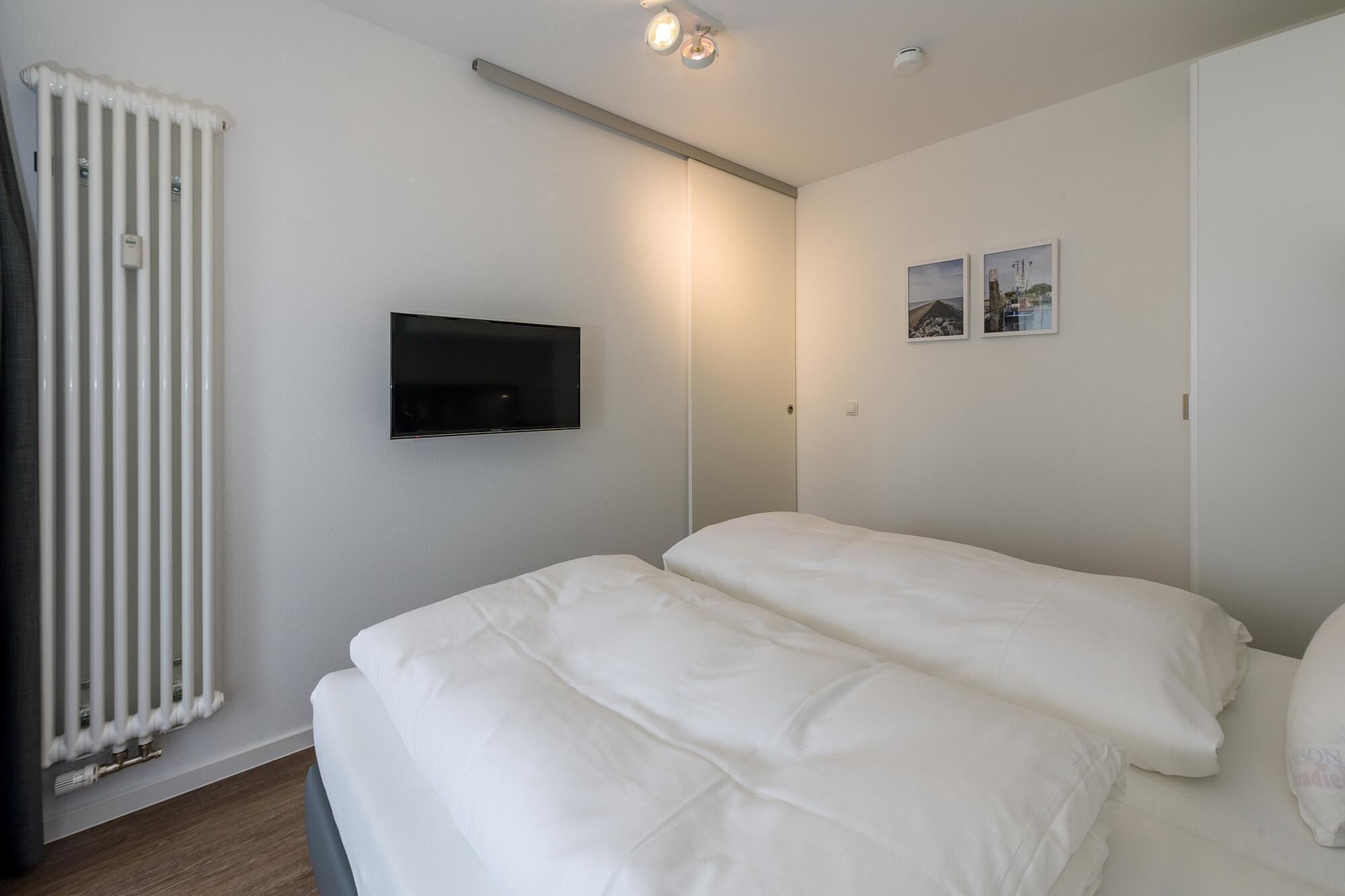 A bedroom with two beds, a radiator, a TV mounted on the wall, and a small window with pictures.