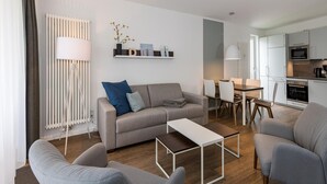 Un salon moderne avec un canapé gris, une table basse et des fauteuils assortis. On y trouve également un coin repas avec une table et des chaises, ainsi qu’une cuisine équipée d’une cuisinière et d’un four.