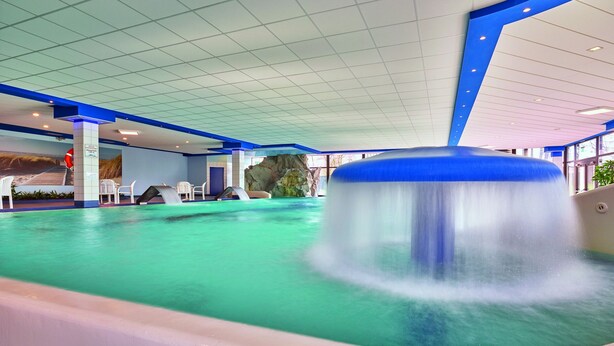 Une piscine intérieure avec un toboggan, un plafond bleu et blanc et des espaces aménagés pour s’asseoir.