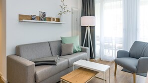 Un salon moderne avec un canapé gris, une table basse blanche et un fauteuil gris. Un radiateur blanc et une lampe sur pied sont visibles sur le mur.