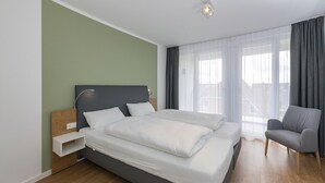 Ein modernes Hotelzimmer mit einem großen Bett, einem Nachttisch, einem Sessel und Blick auf die Stadt durch große Fenster.
