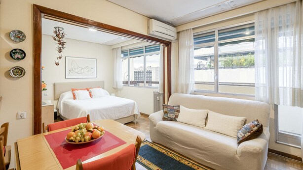 A hotel room with a bed, a sofa, a dining table with fruit, and a view of the city through large windows.