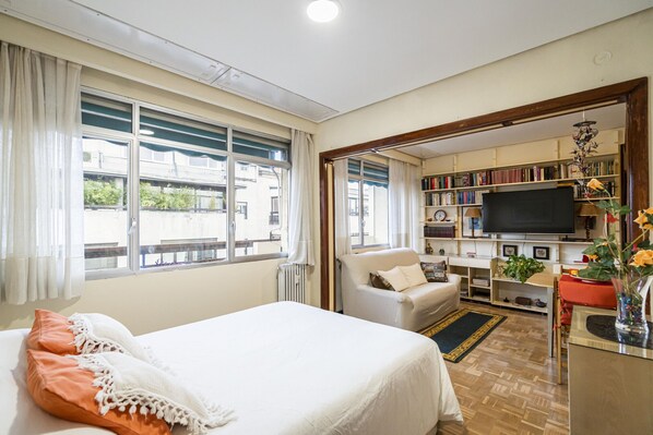A hotel room with a bed, a sofa, a TV, a bookshelf, and a view of the outside.
