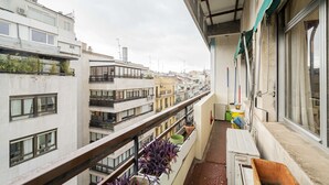A narrow balcony with potted plants, a window, and a view of adjacent buildings.