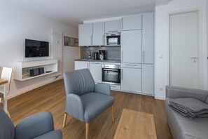 Een moderne keuken met witte kastjes, een inbouwoven en een koffiezetapparaat. In de woonkamer staat een grijze fauteuil en een grijze bank.