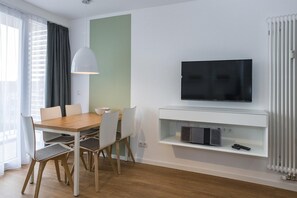 Een moderne woonkamer met een houten eettafel, witte stoelen, een flatscreen televisie aan de muur en een inbouwplank onder de televisie.
