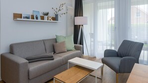 A modern living room with a grey sofa, a matching armchair, a wooden coffee table, and a floor lamp. A shelf with decorative items is mounted on the wall.
