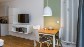 Un espace de vie moderne et compact, avec un téléviseur posé sur un meuble blanc, une table à manger avec des chaises, un luminaire suspendu et un radiateur.