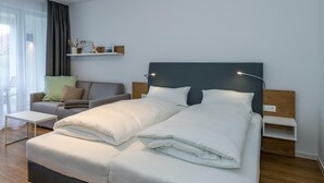 Une chambre d’hôtel moderne avec un lit double, une petite table de chevet, une commode, un canapé et une étagère murale agrémentée d’objets décoratifs.