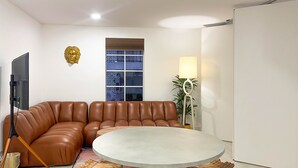 A modern living room with a brown leather sofa, a round table with wooden stools, a TV on a stand, and a window with a view of a building.