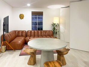 A modern living room with a brown leather sofa, a round table with wooden stools, a TV on a stand, and a window with a view of a building.