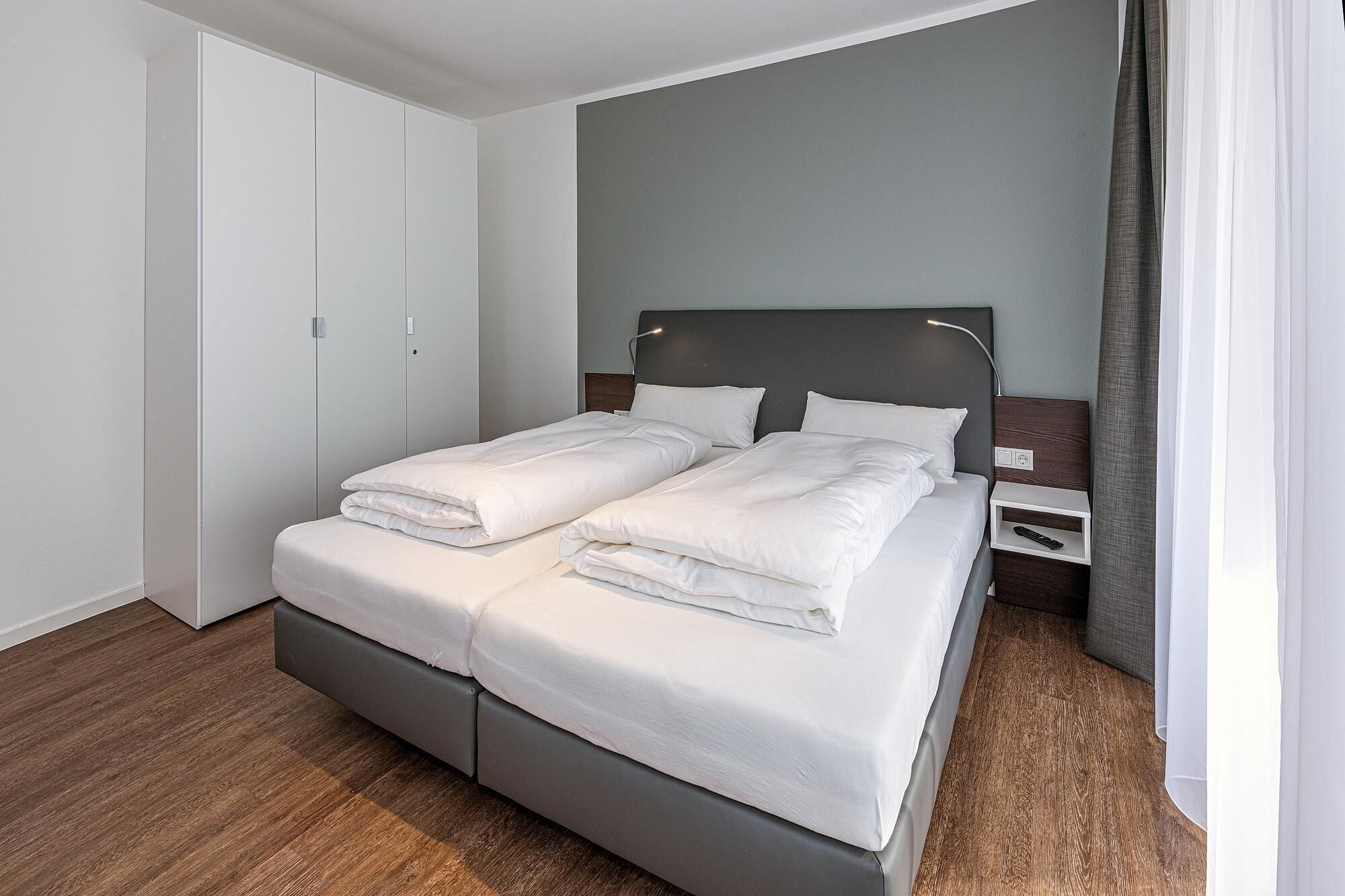 A neatly made bed with white linens, a grey headboard, and a bedside table with a remote control.