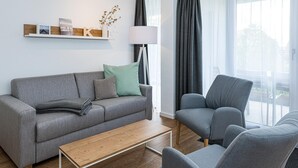 Una sala de estar moderna con un sofá gris, un sillón a juego, una mesa de centro de madera y una lámpara de pie. En la pared, hay un estante con fotos enmarcadas y un jarrón con flores.