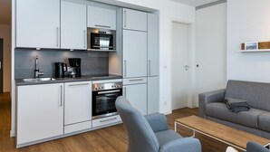 Una cocina moderna con armarios blancos, horno integrado y cafetera. En la sala de estar hay un sofá gris y una mesa de madera.
