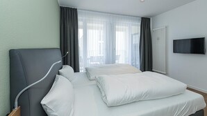 A modern hotel room with a large bed, a flat-screen TV, and a view of the outdoors through large windows. - Strandnahes Ferienapartment mit Balkon, Parkplatz, Schwimmbad- & Saunanutzung (Nordseeheilbad Bensersiel)