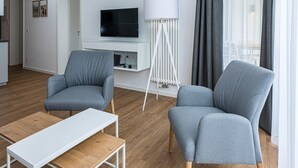 A modern living room with two grey armchairs, a white coffee table, a flat-screen TV on a white stand, and a floor lamp. - Strandnahes Ferienapartment mit Balkon, Parkplatz, Schwimmbad- & Saunanutzung (Nordseeheilbad Bensersiel)