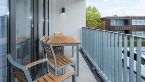 A balcony with a wooden table and chairs, a metal railing, and a view of buildings and trees. - Strandnahes Ferienapartment mit Balkon, Parkplatz, Schwimmbad- & Saunanutzung (Nordseeheilbad Bensersiel)