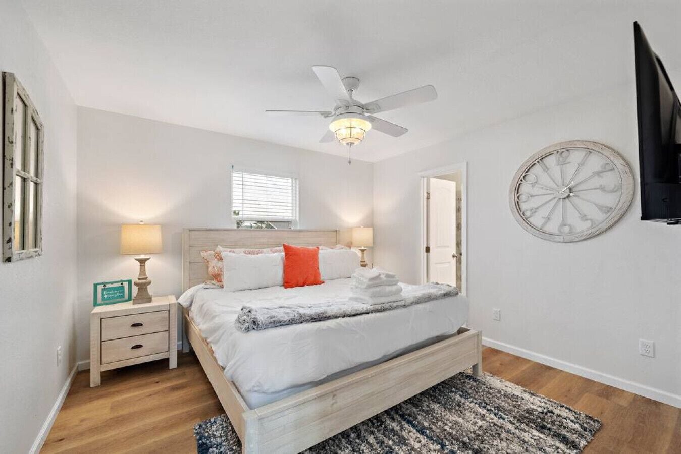 A bedroom with a bed, bedside lamps, a ceiling fan, a clock on the wall, and a window.