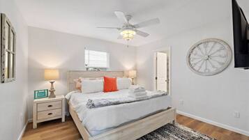 A bedroom with a bed, bedside lamps, a ceiling fan, a clock on the wall, and a window.