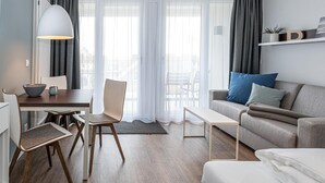 A modern living room with a dining area, a sofa, a coffee table, and a chair. There is a large window with sheer curtains and a view of the outdoors.