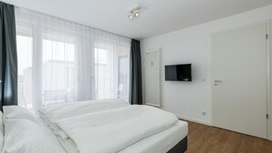 A modern bedroom with a bed, a TV mounted on the wall, and a window with sheer curtains.