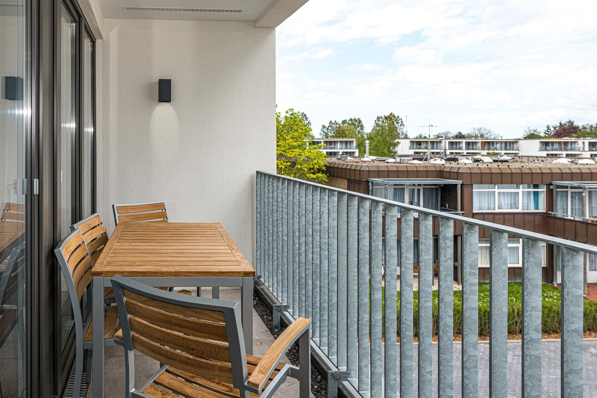 Ein Balkon mit Holztisch und -stühlen, einem Metallgeländer und Blick auf Wohnhäuser im Hintergrund.