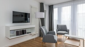A modern living room with a flat-screen TV, a white entertainment unit, two grey armchairs, a small white table, and a floor lamp.