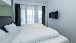 A hotel room with a bed, a TV mounted on the wall, and a window with curtains. - Strandnahes Ferienapartment mit Balkon, Parkplatz, Schwimmbad- & Saunanutzung (Nordseeheilbad Bensersiel)