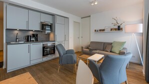 A compact living space with a kitchenette, a sofa, a coffee table, and a floor lamp. - Strandnahes Ferienapartment mit Balkon, Parkplatz, Schwimmbad- & Saunanutzung (Nordseeheilbad Bensersiel)