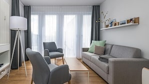 Un salón moderno con un sofá gris, dos sillones, una mesa de centro y una lámpara. Cuenta con un ventanal con cortinas transparentes y vistas al exterior.