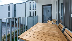 Un balcón con mesa y sillas de madera, barandilla de metal y vistas a edificios modernos.