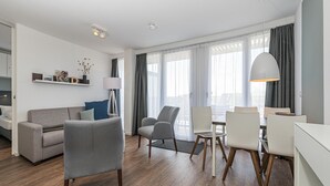 Un salon moderne avec un canapé gris, deux fauteuils, une table à manger avec des chaises et une grande fenêtre avec des rideaux.