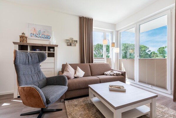 A modern living room with a sofa, armchair, and coffee table. There is a large window with blinds, a lamp, and a picture frame on the wall.
