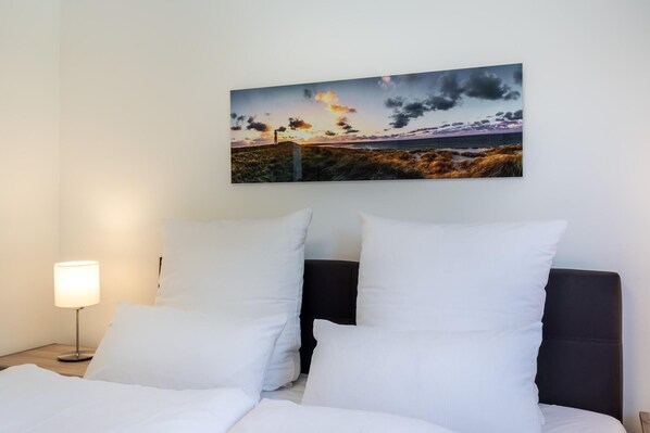 A hotel room with a bed, white pillows, a nightstand with a lamp, and a painting of a sunset over the sea on the wall.