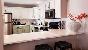 Una cocina moderna con gabinetes blancos, un refrigerador de acero inoxidable, un microondas negro y una estufa. Hay dos taburetes negros y un florero decorativo sobre la encimera.
