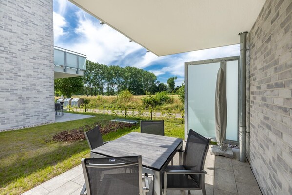 Un patio exterior con una mesa y sillas, un ventanal de vidrio y vistas a un jardín y árboles.