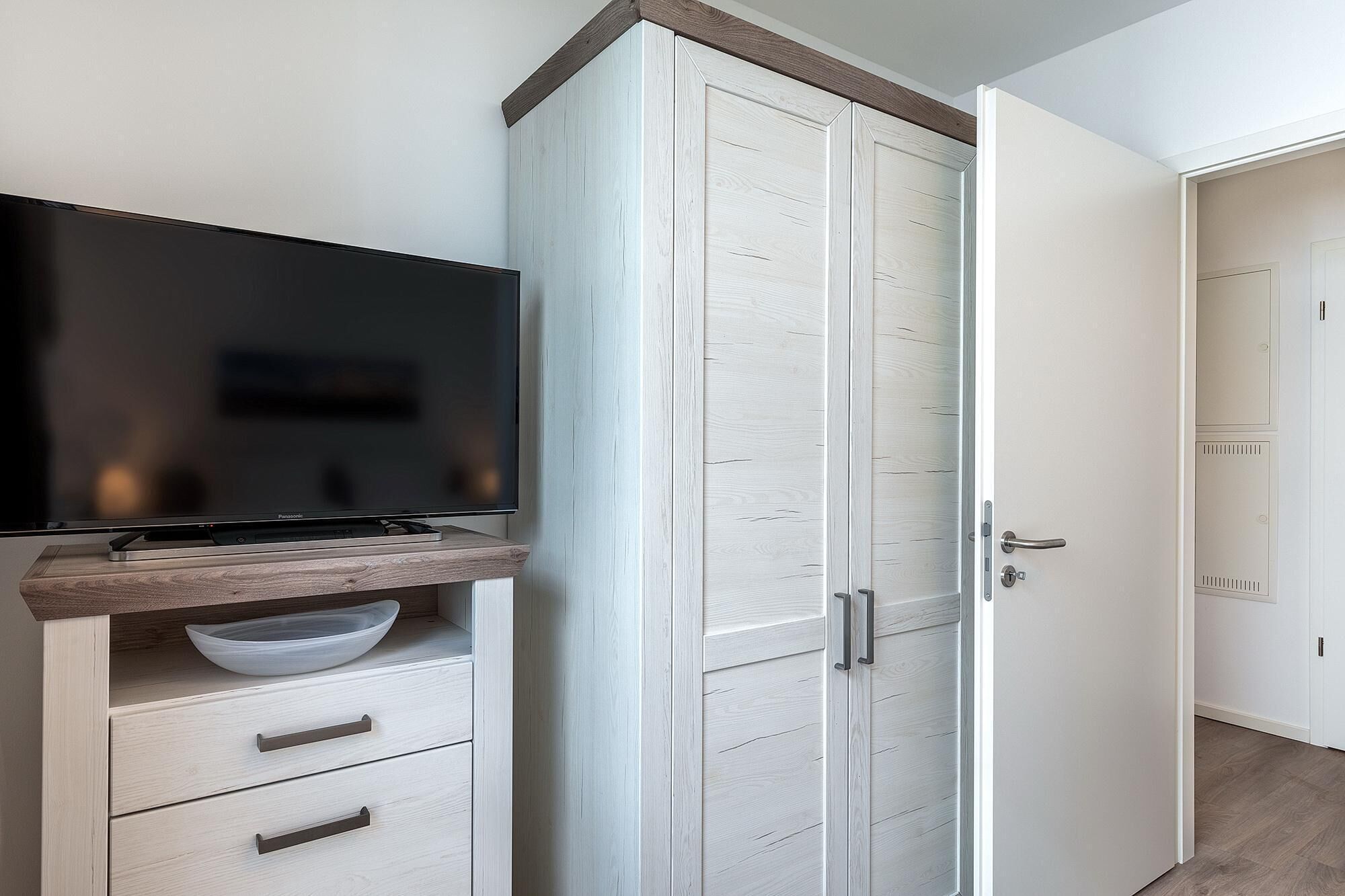 Una habitación con una televisión sobre un estante de madera, un armario blanco con puerta, y una puerta con manija.