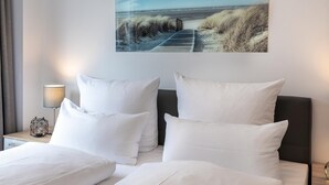 A hotel room with two beds, white bedding, a nightstand with a lamp, and a beach scene painting on the wall.
