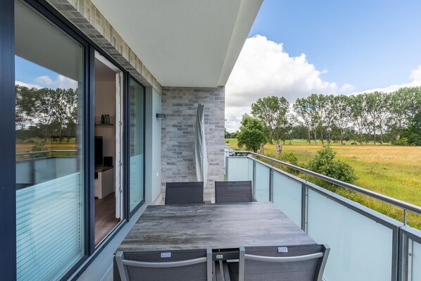 A balcony with a table and chairs, a view of a grassy field, and a brick wall. - Tolles Familien-apartment mit Balkon; Spielplatz und Saunabereich in der Anlage! (Ostseebad Boltenhagen)