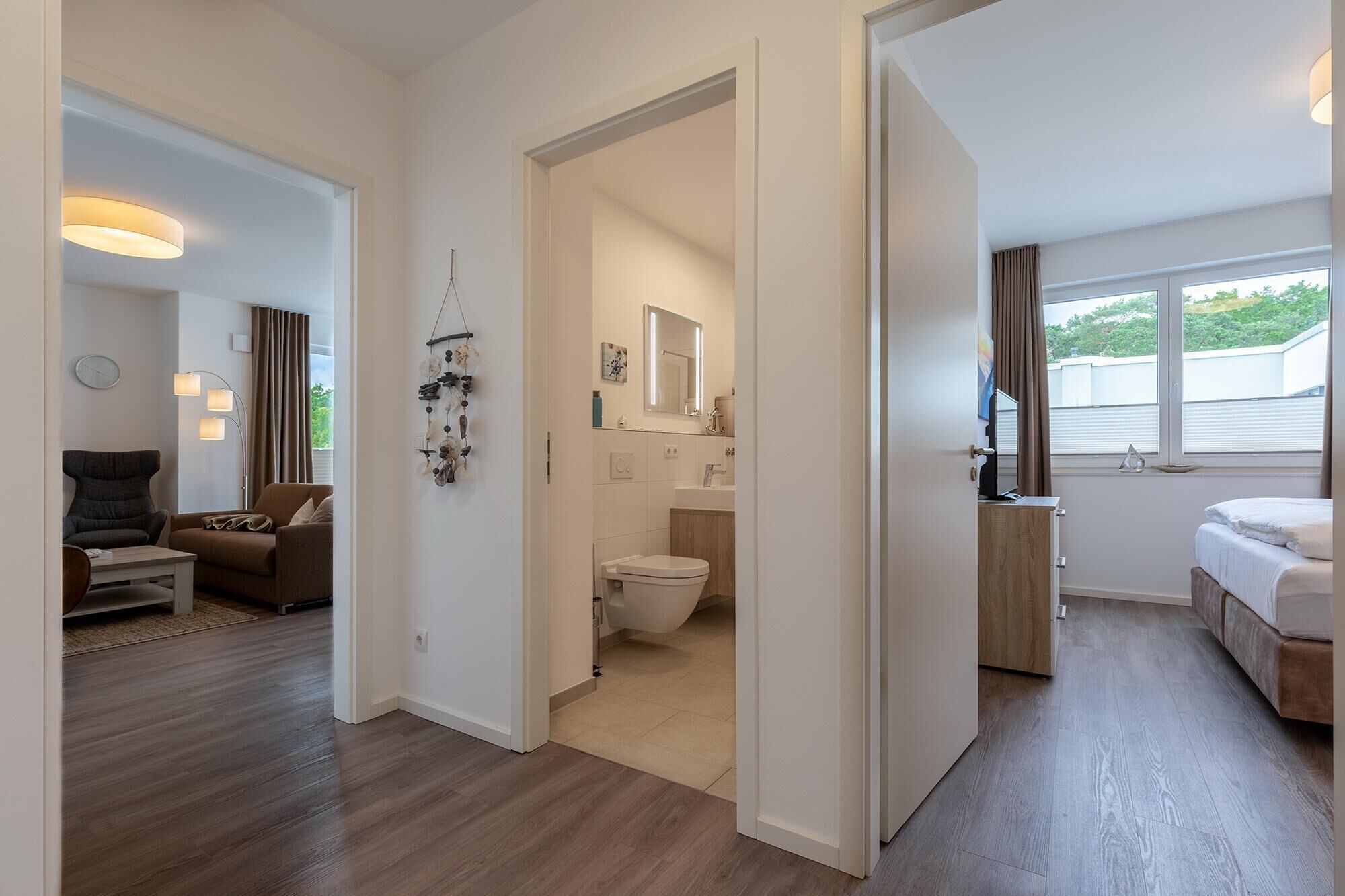 Una habitación de hotel moderna con piso de madera, una cama, un escritorio y una silla. Incluye un baño con inodoro, lavamanos y espejo. La habitación tiene un ventanal con vista a follaje exterior.