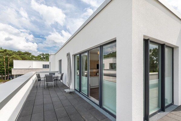 A modern building with a patio area featuring a table and chairs, large sliding glass doors, and a view of the surrounding area.