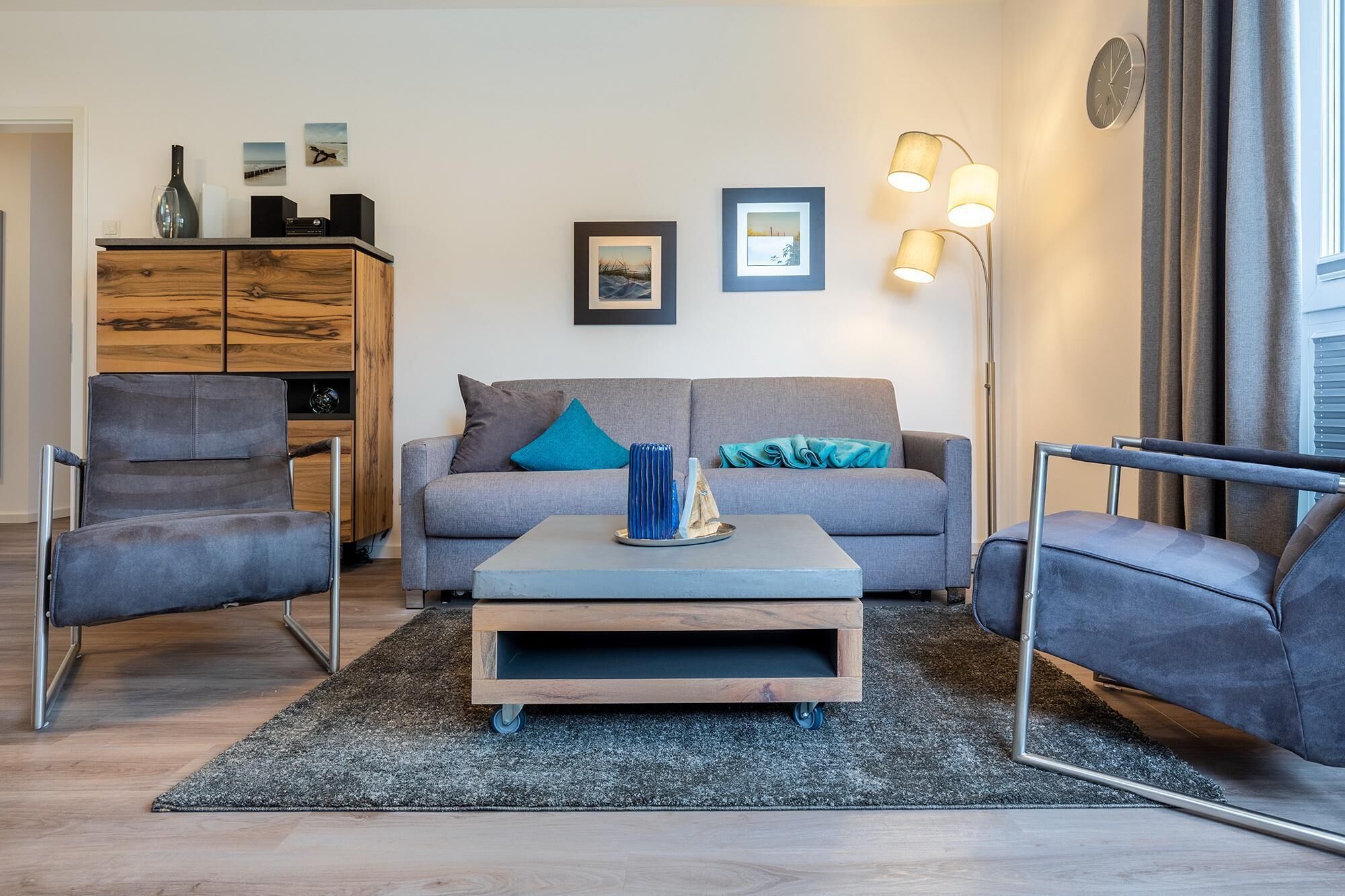 Uma sala de estar moderna com um sofá cinza, duas poltronas, uma mesa de centro e um aparador de madeira. Há quadros nas paredes e um abajur de chão.