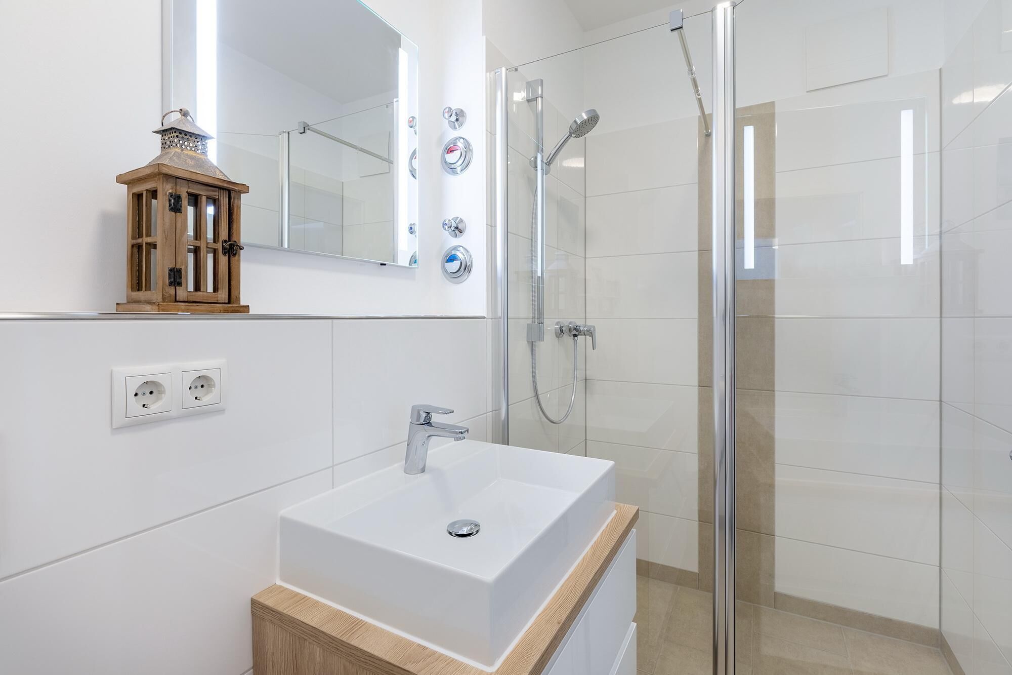 Une salle de bain moderne avec un lavabo blanc, un meuble en bois, une cabine de douche en verre et un miroir avec éclairage intégré.